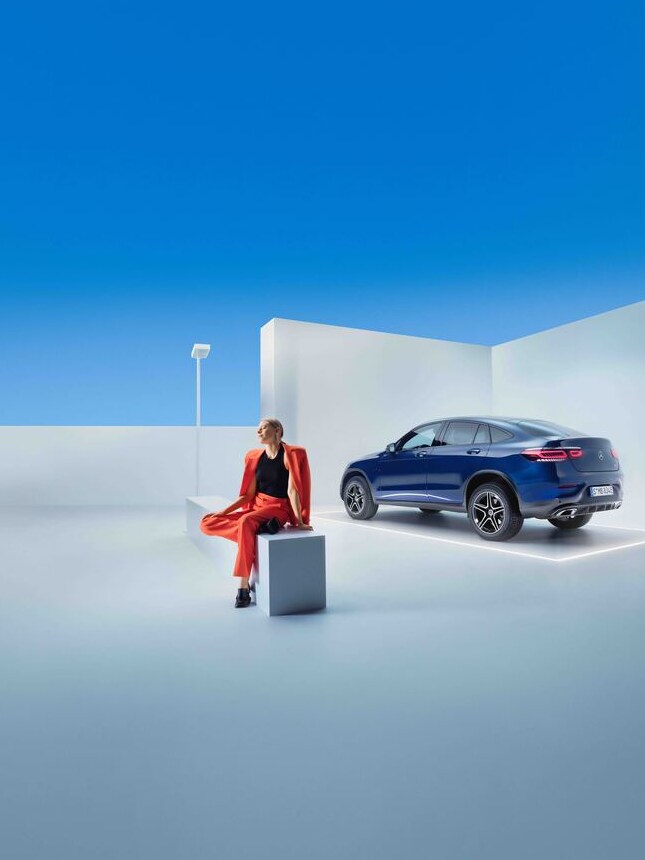 Kobieta siedzi na ławce obok ciemnoniebieskiego Mercedesa GLC.