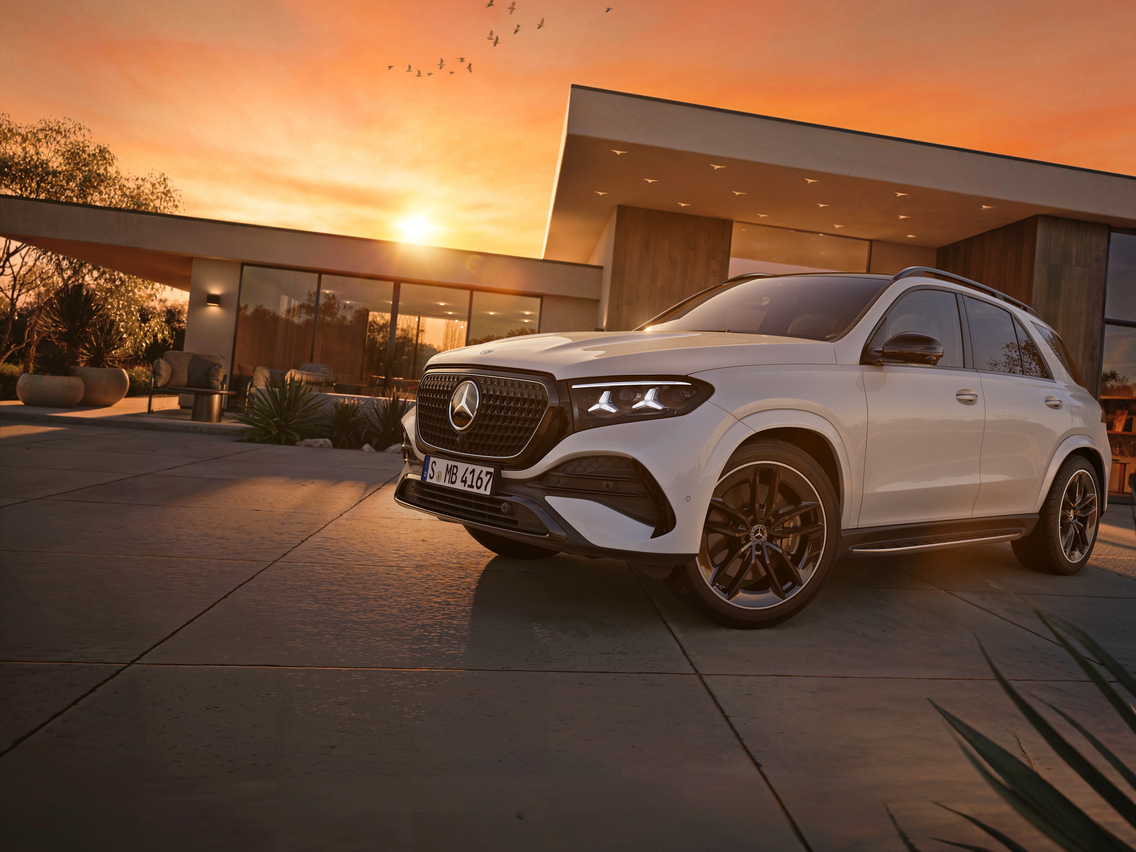 Biały Mercedes-Benz GLE SUV zaparkowany przed domem na tle zachodzącego słońca.