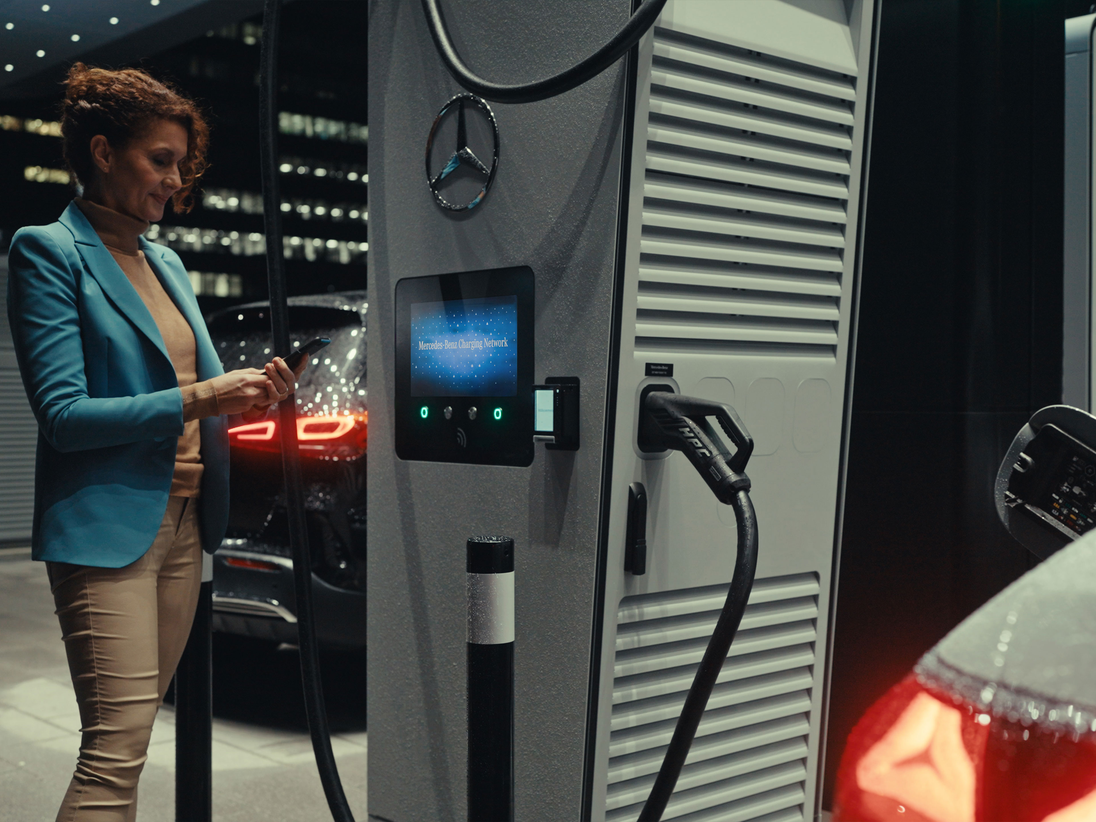 Aplikacja Mercedes-Benz | Ładowanie w drodze | Mercedes-Benz Kobieta trzymająca w dłoni smartfona, która stoi obok stacji ładowania.