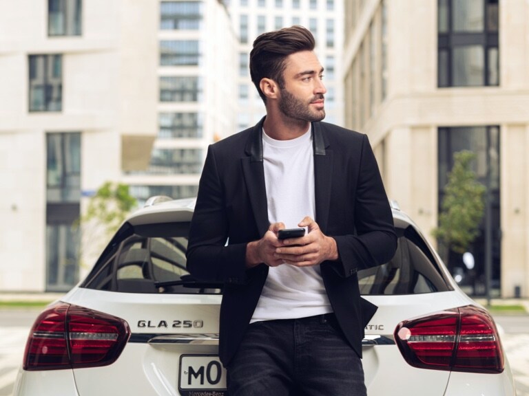 Mężczyzna opiera się o swojego Mercedesa GLA i trzyma w ręku smartfona.