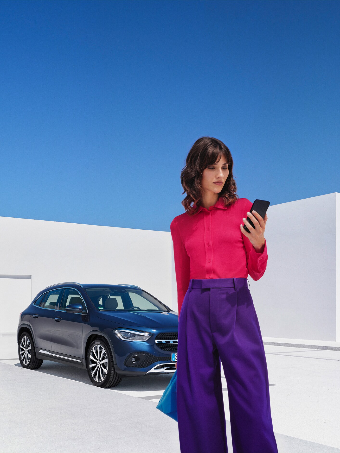 Kobieta patrzy na smartfona na tle niebieskiego Mercedesa GLA.