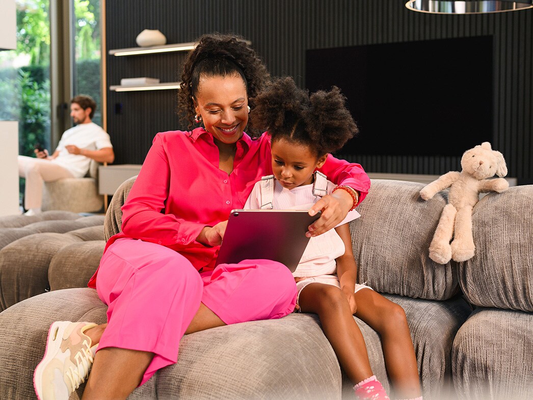Osoba i dziecko siedzące na kanapie i korzystające z urządzenia mobilnego, z inną osobą siedzącą w oddali.