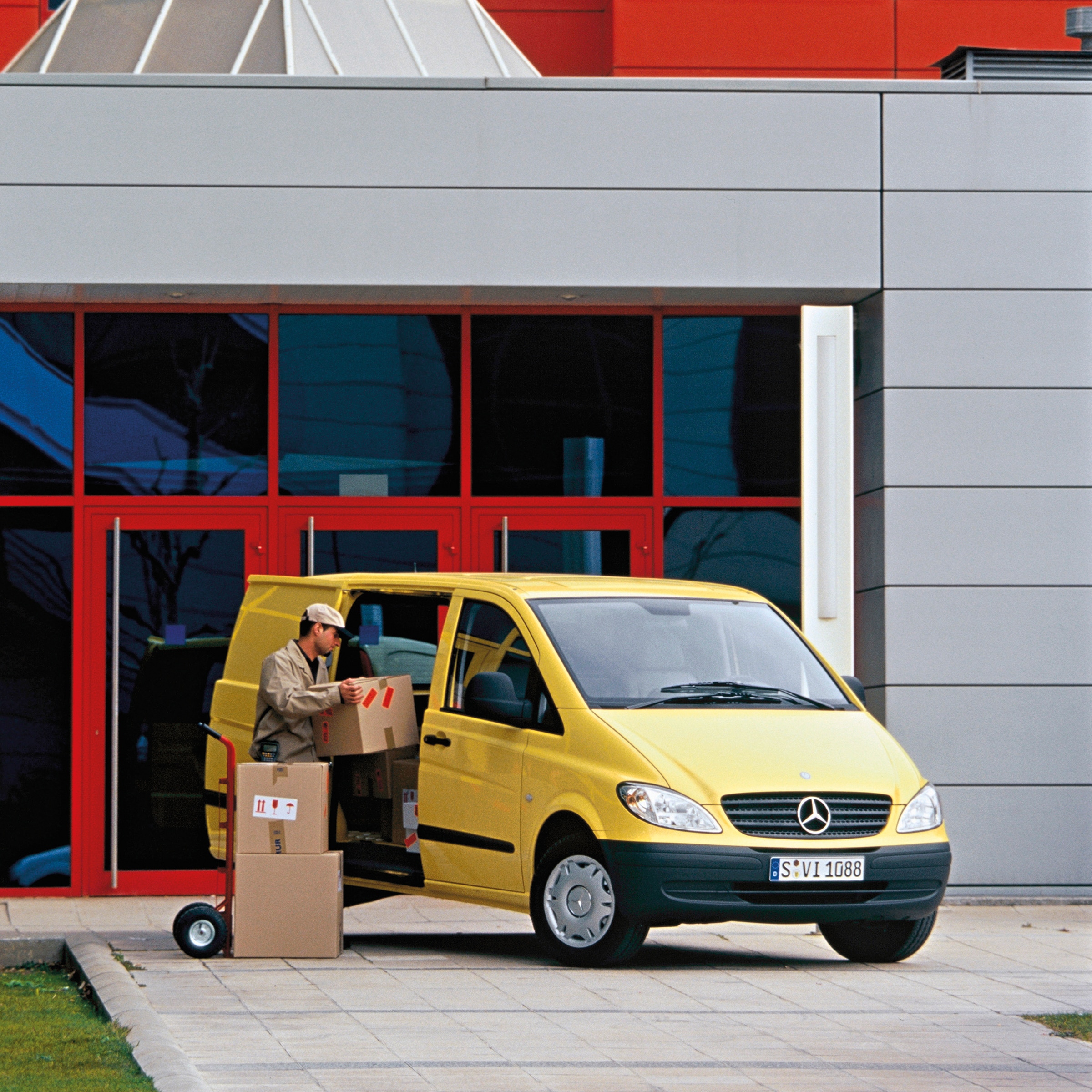 Druga generacja Mercedes-Benz Vito | Historia pojazdów użytkowych | Mercedes-Benz Żółty Mercedes-Benz Vito zaparkowany przed budynkiem i pracownik rozładowujący paczki