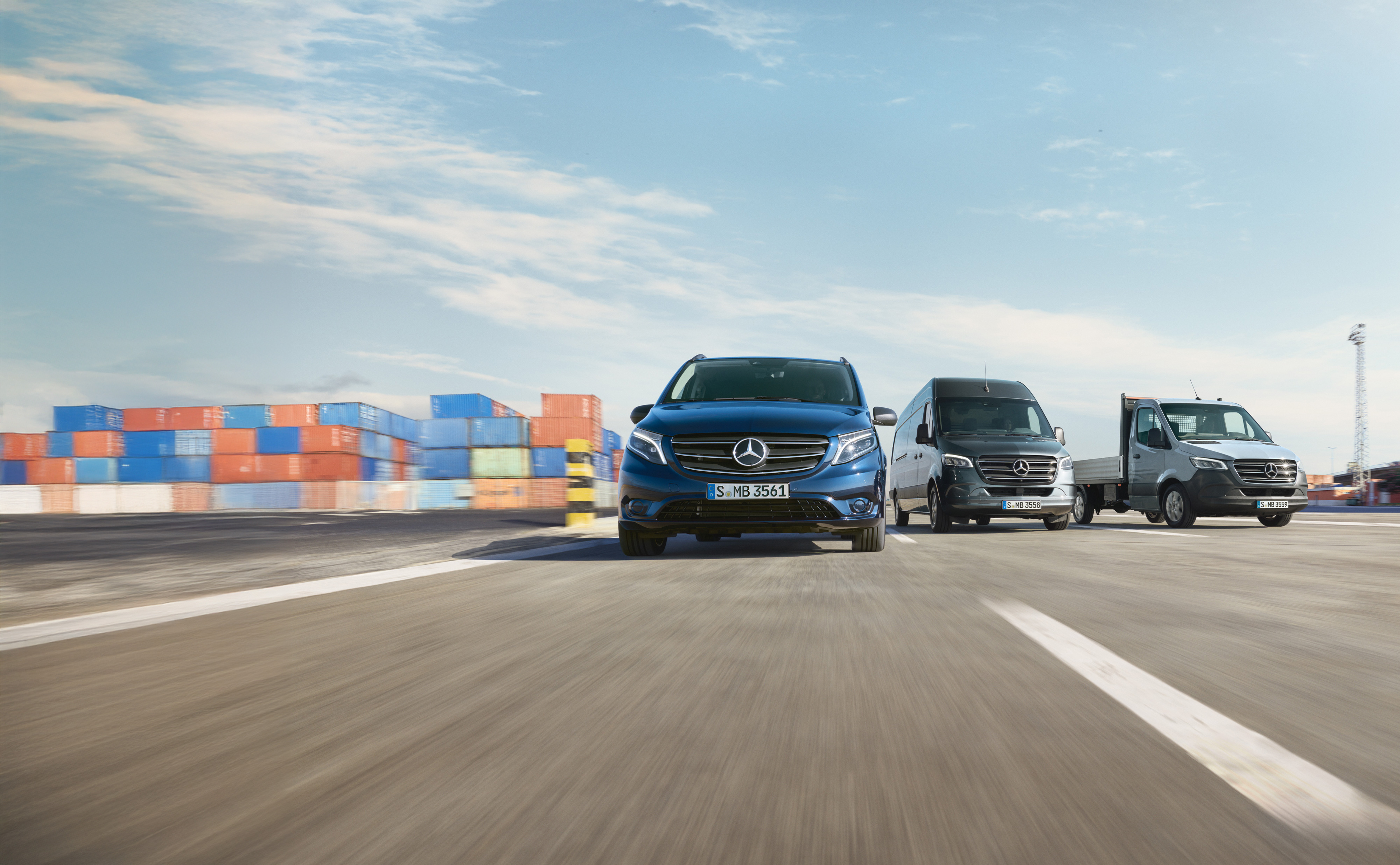 Service24h | Samochody dostawcze Mercedes-Benz Szereg samochodów dostawczych Mercedes-Benz jadących obok siebie na drodze obok doku przeładunkowego.