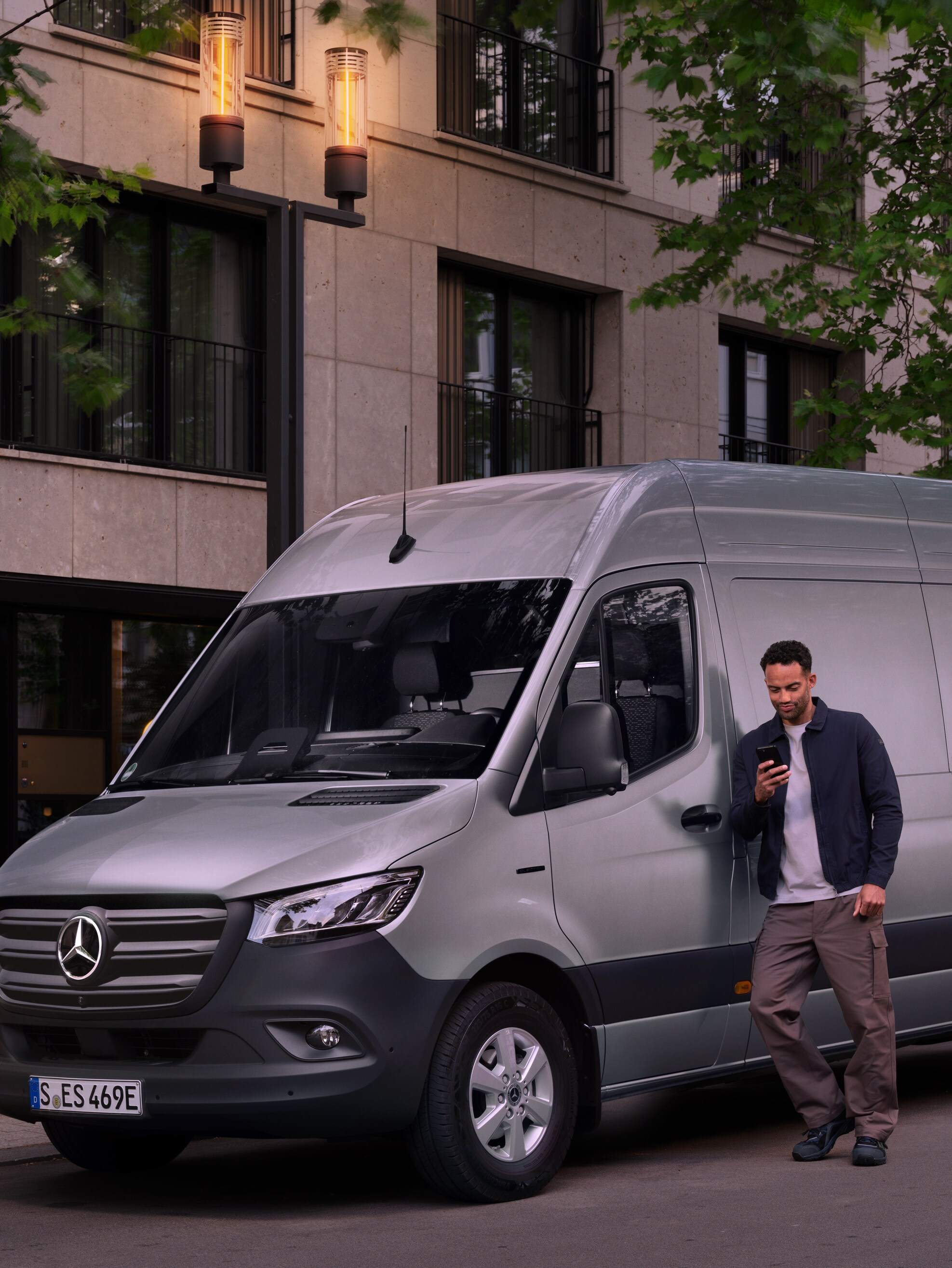 Mężczyzna opierający się o srebrny samochód dostawczy Mercedes-Benz Sprinter i korzystający ze smartfona.