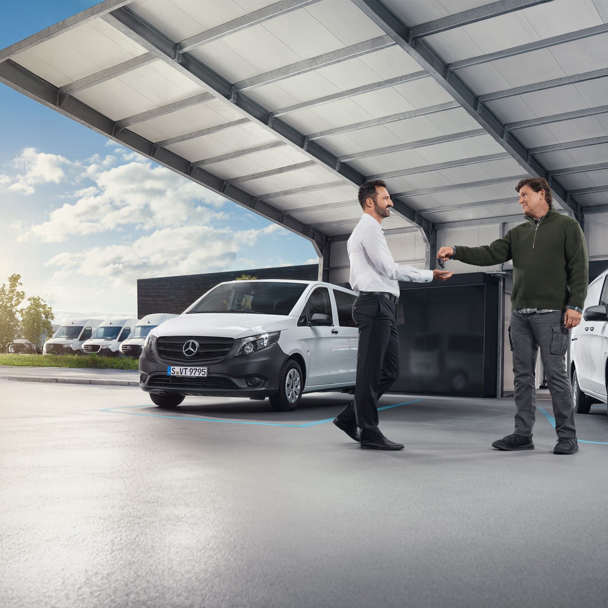 Dwóch mężczyzn rozmawia przed szeregiem białych samochodów dostawczych Mercedes-Benz w hali.