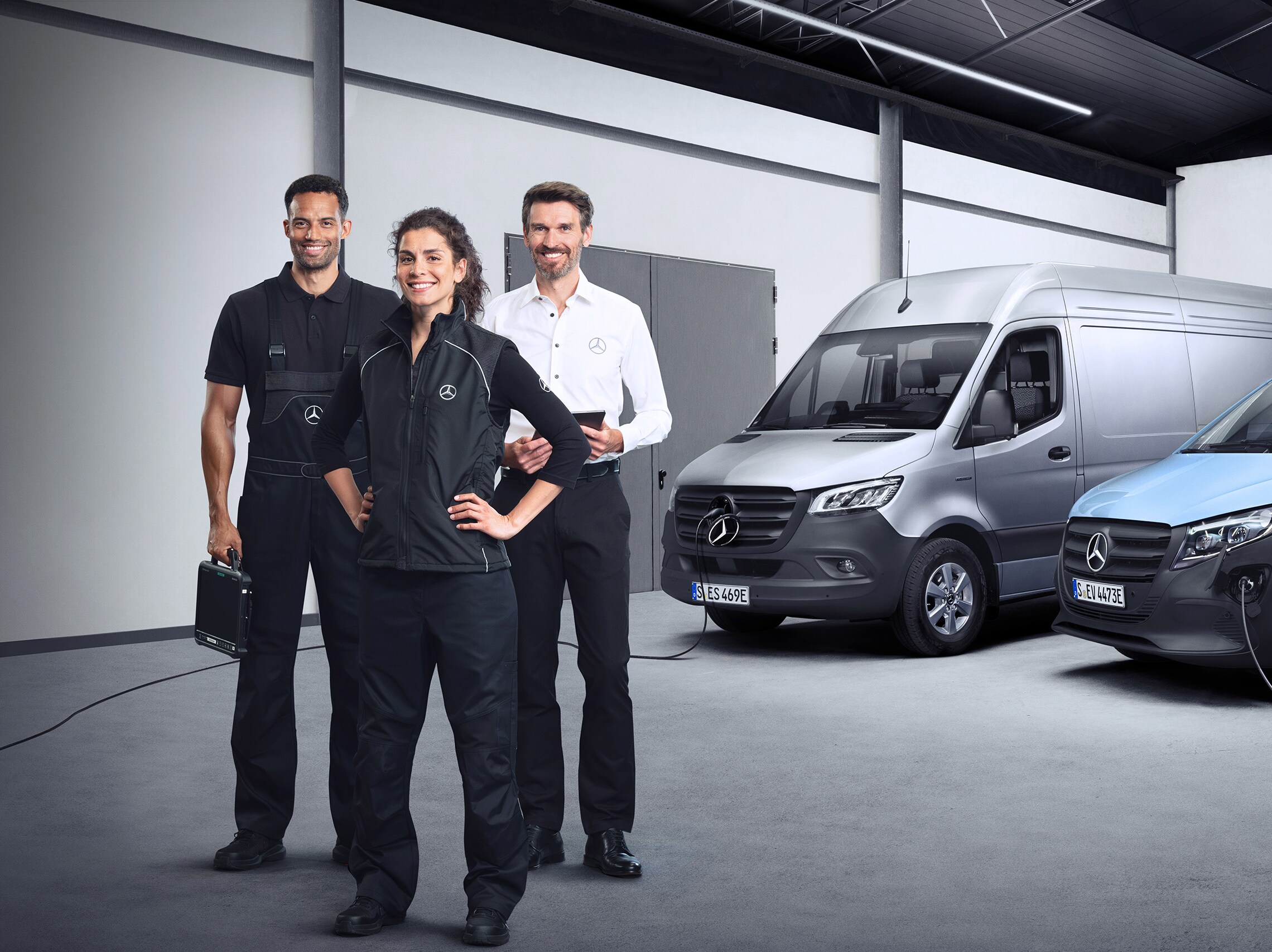 Trzej pracownicy z zespołu serwisowego Mercedes-Benz stoją przed samochodami Mercedes-Benz eSprinter i Mercedes-Benz eVito.