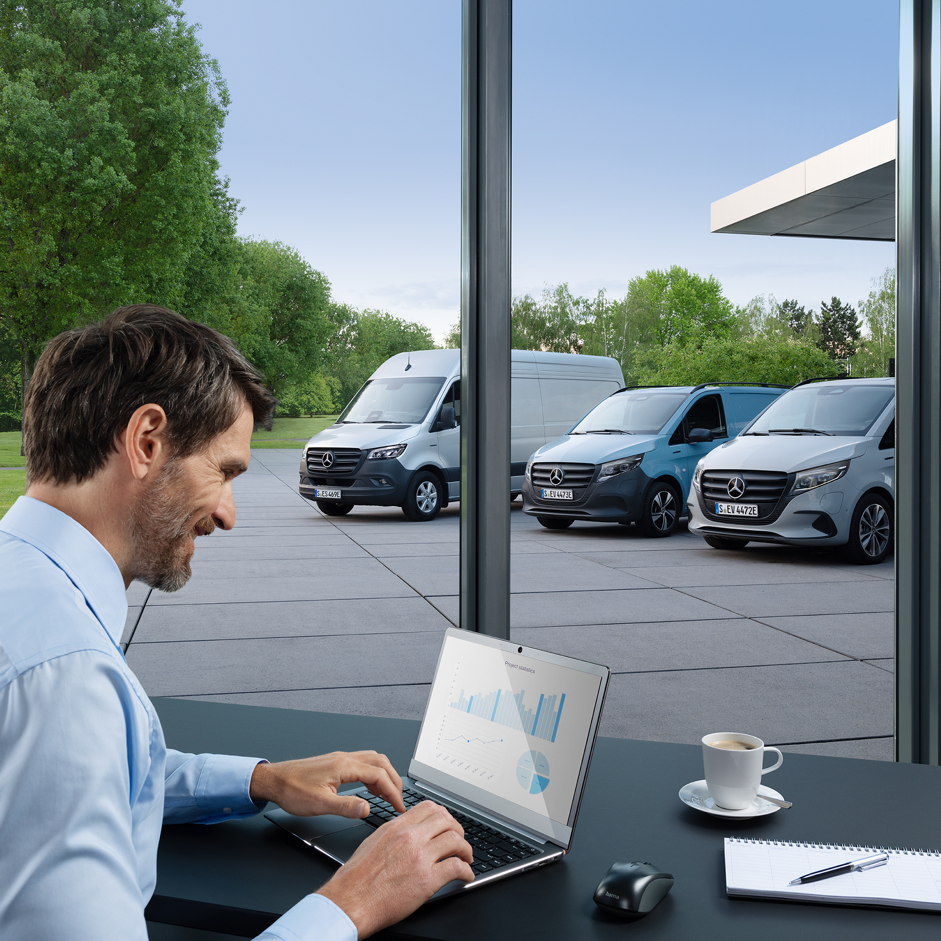 Pojazdy elektryczne Mercedes-Benz | Umowy serwisowe Mercedes-Benz Osoba w biurze siedzi przed ekranem laptopa. Trzy elektryczne samochody dostawcze Mercedes-Benz zaparkowane na zewnątrz.