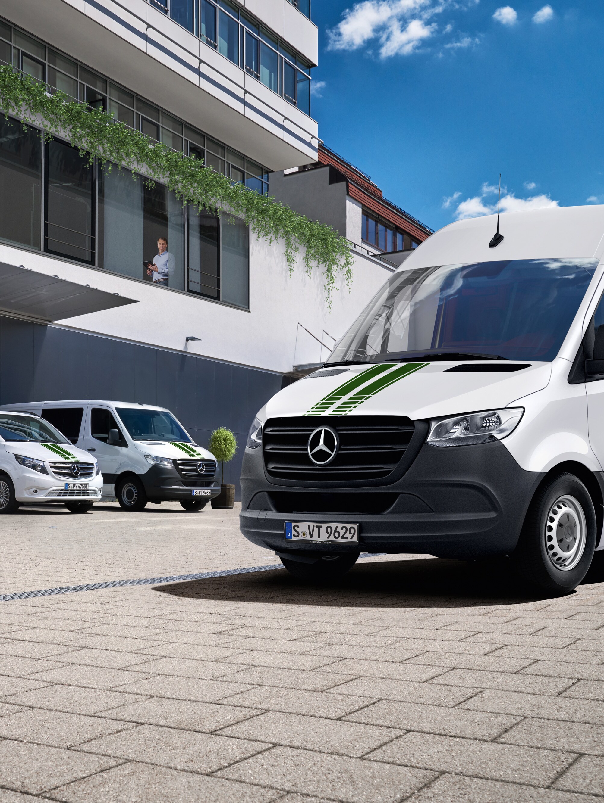 Flota białych samochodów dostawczych Mercedes-Benz przed budynkiem biurowym, w słoneczny dzień, na tle błękitnego nieba.