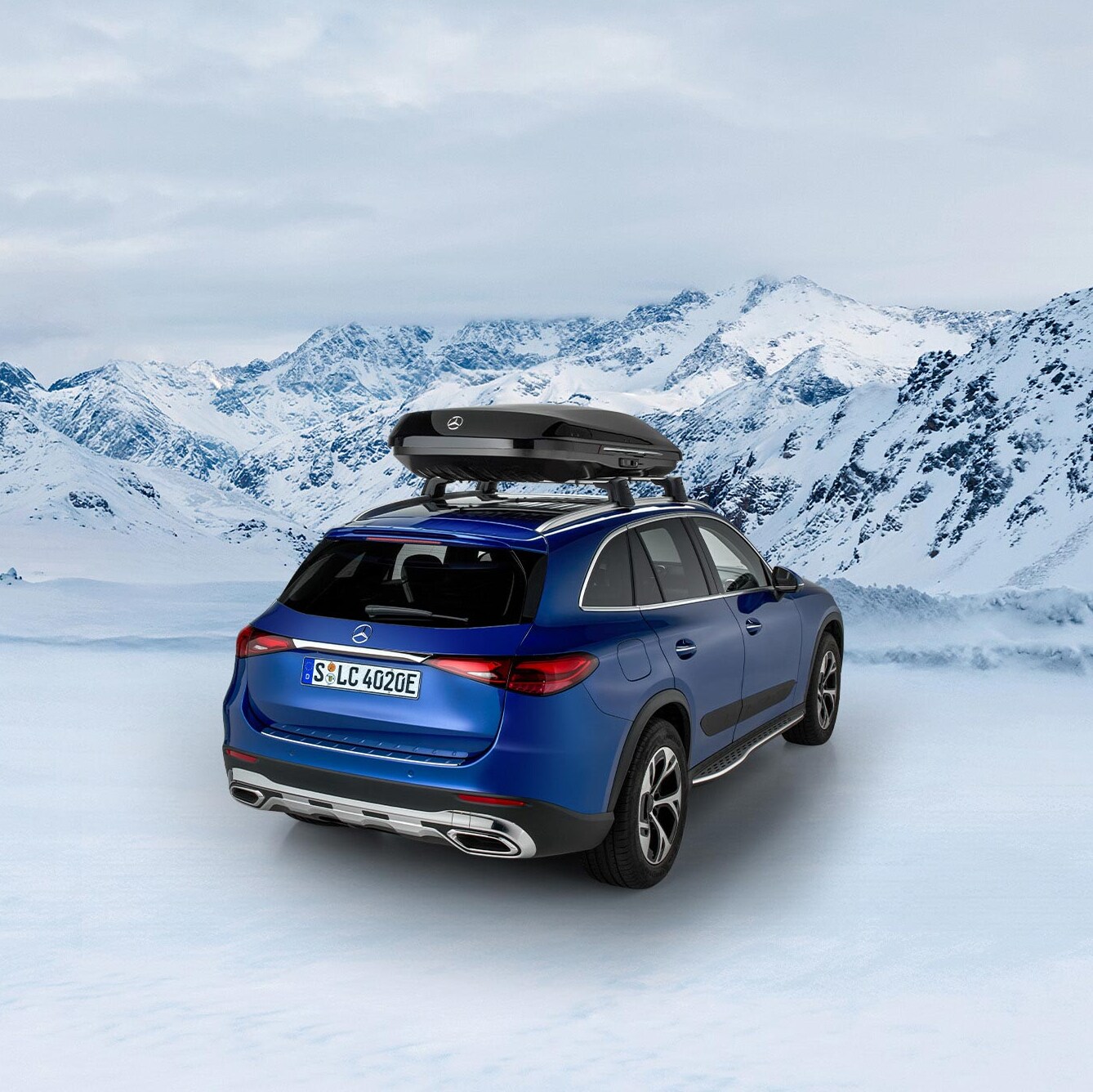 Niebieski Mercedes GLC SUV z boksem dachowym stoi zaparkowany na tle ośnieżonych gór.