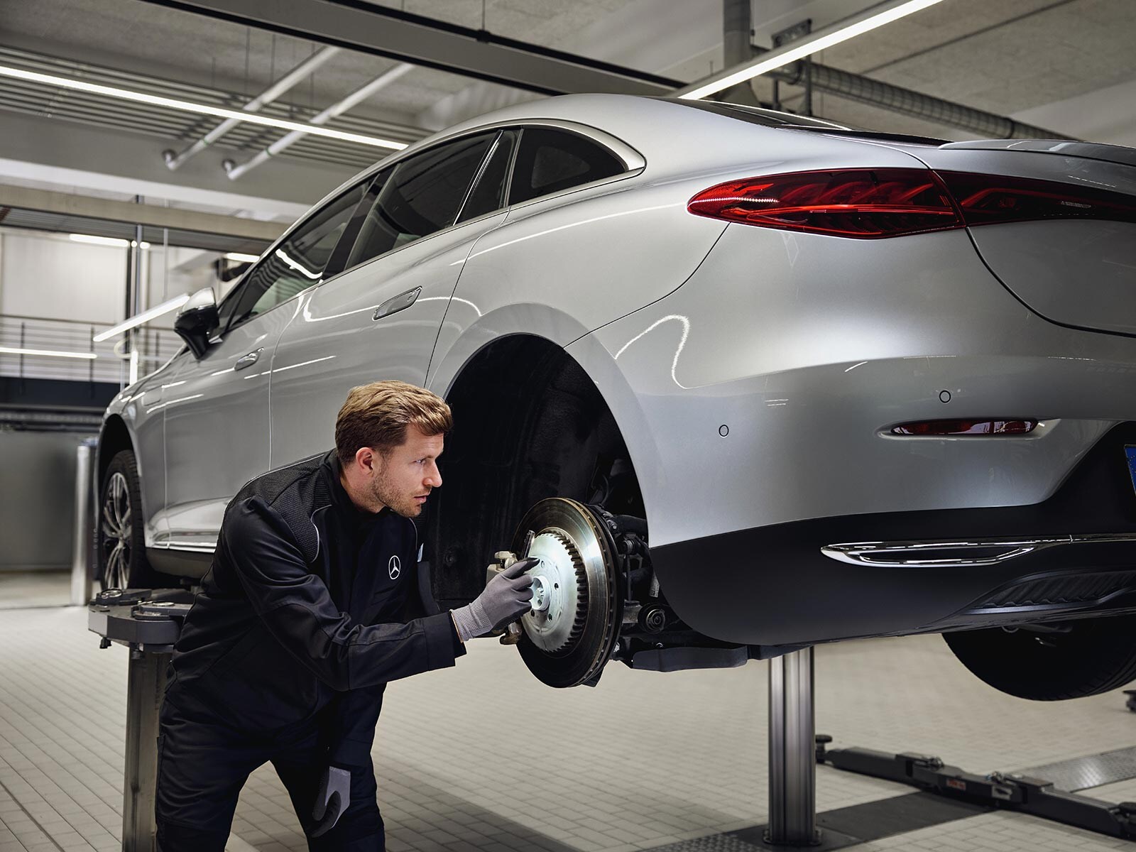 Samochód Mercedes-Benz ustawiony na podnośniku, a technik sprawdza tylny układ hamulcowy w ramach serwisu wymiany oryginalnych części.