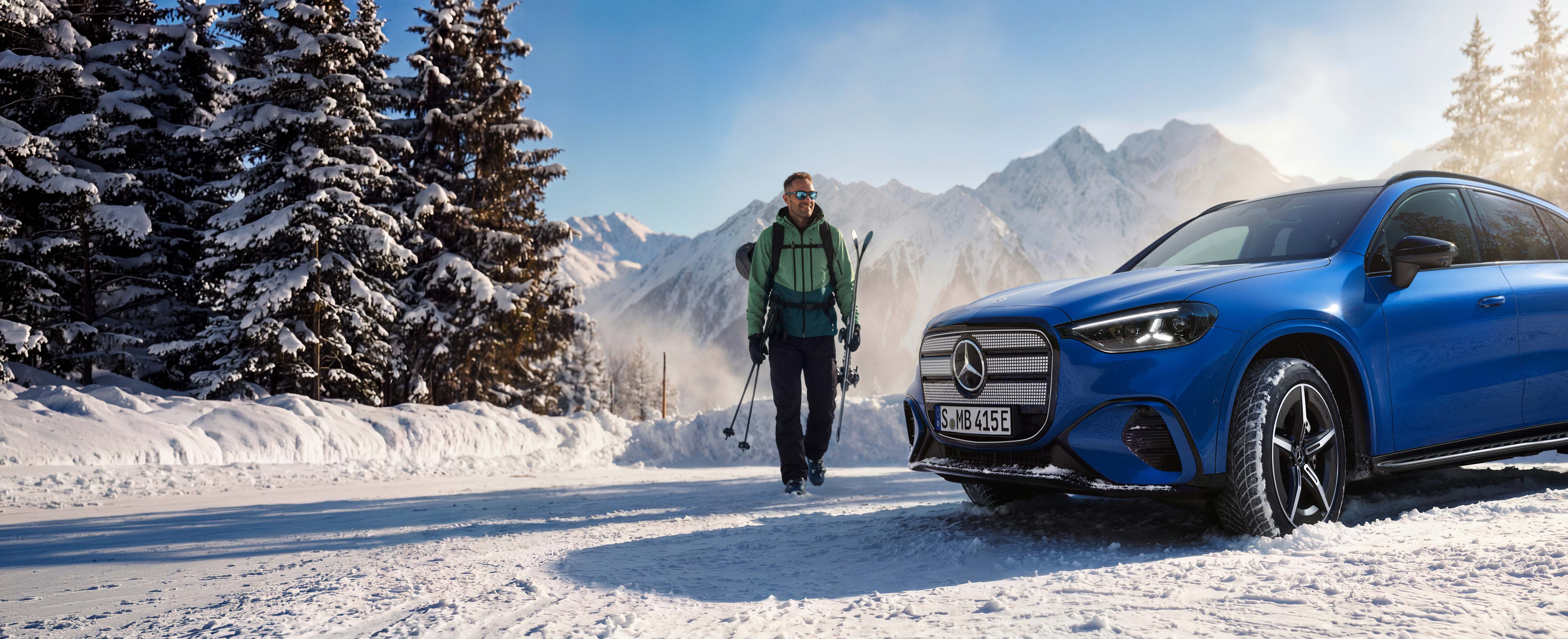 Oferty specjalne | Mercedes-Benz Uśmiechnięty mężczyzna niosący narty podchodzi do błękitnego Mercedesa stojącego na ośnieżonym parkingu.