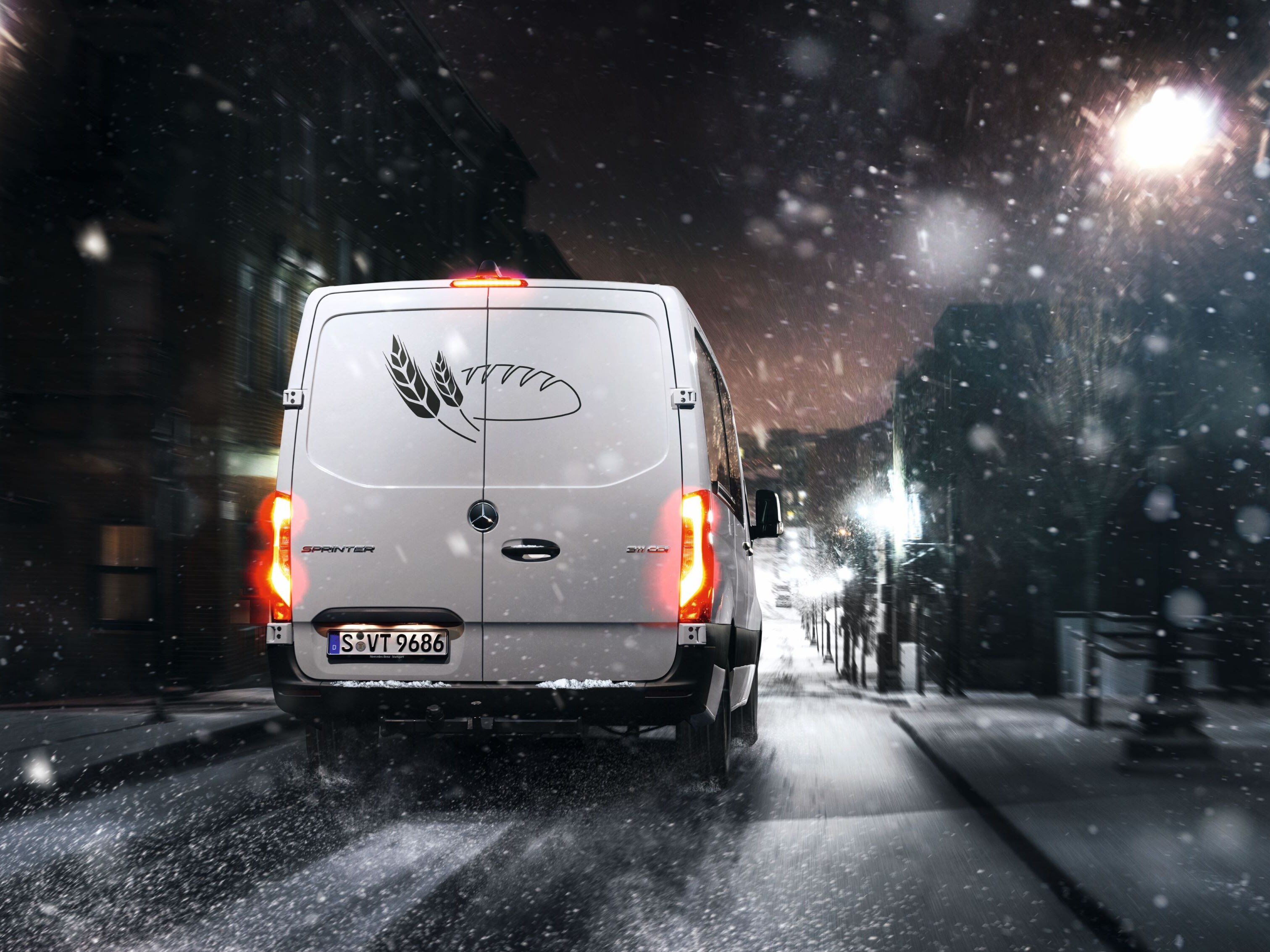 Mercedes-Benz Sprinter z logo piekarni porusza się wśród padającego śniegu po oświetlonej latarniami ulicy.