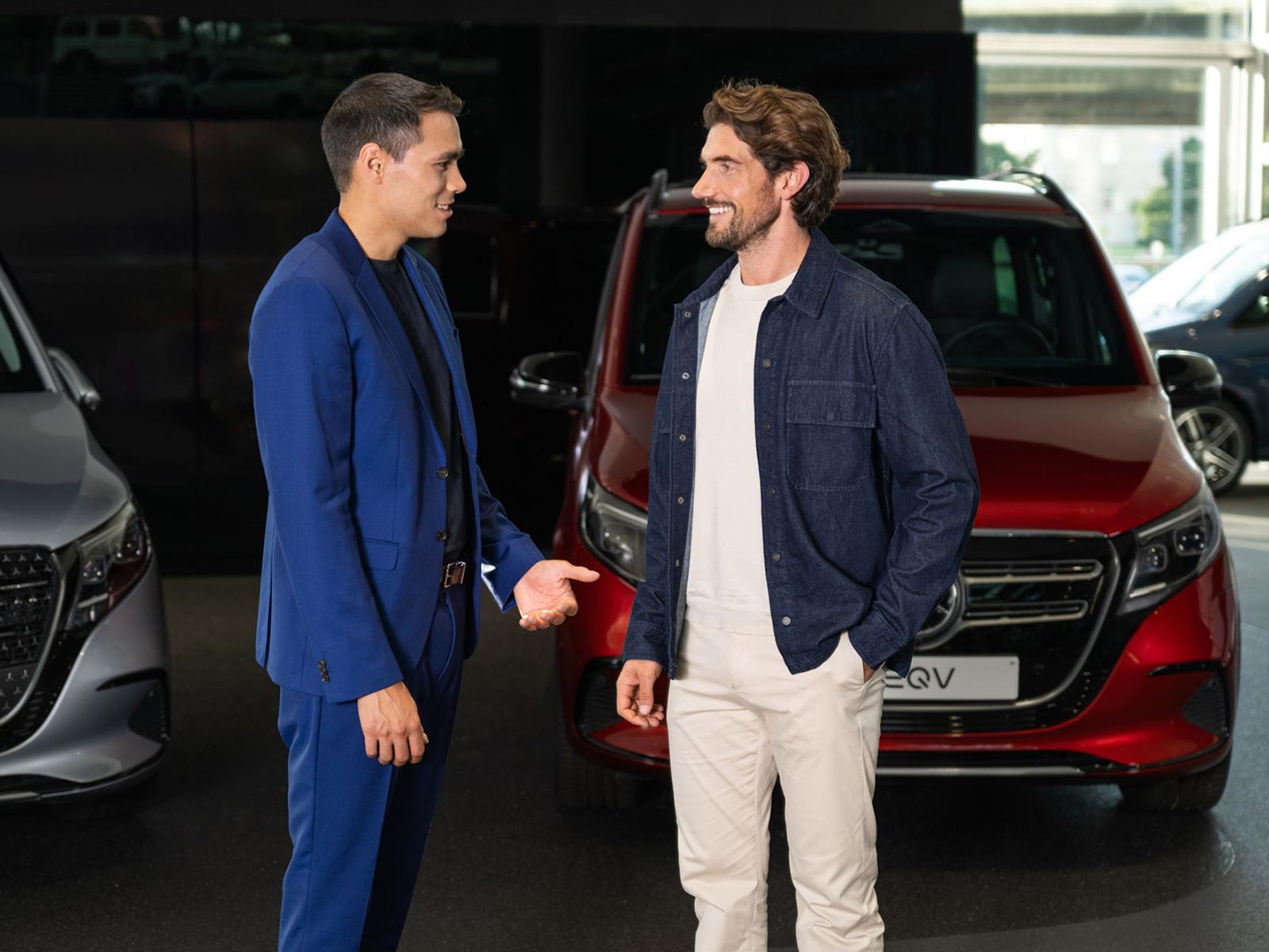 Oferta finansowania dla osób prywatnych | EQV | Mercedes-Benz Dwóch mężczyzn rozmawia stojąc obok czerwonego Mercedesa EQV.