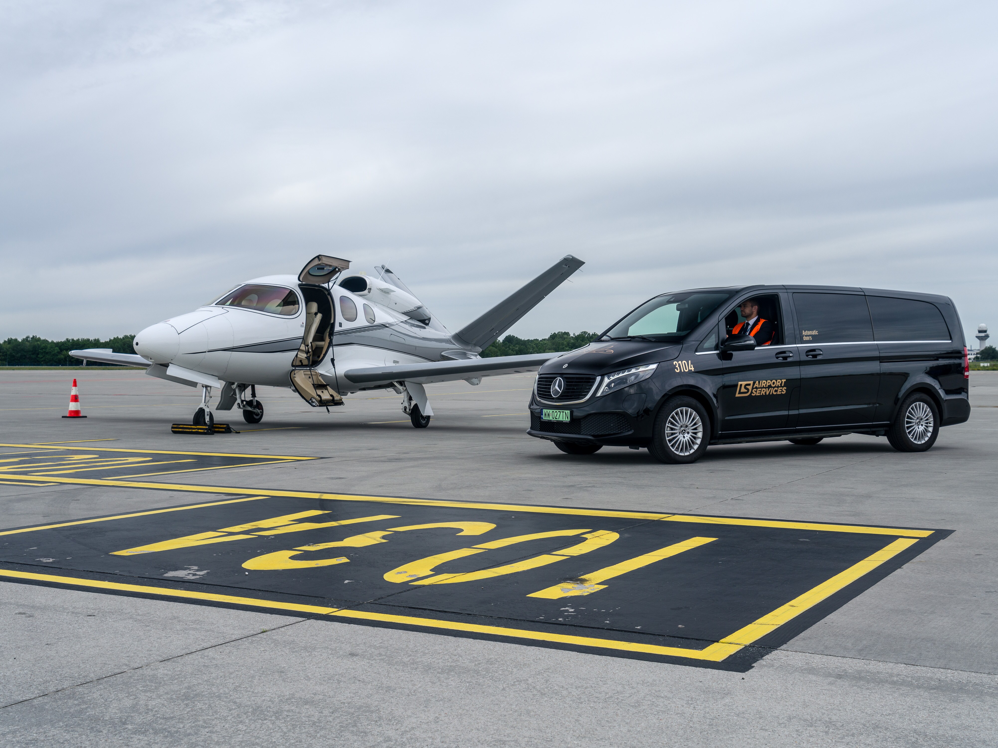 Czarny Mercedes EQV na płycie lotniska obok prywatnego odrzutowca.