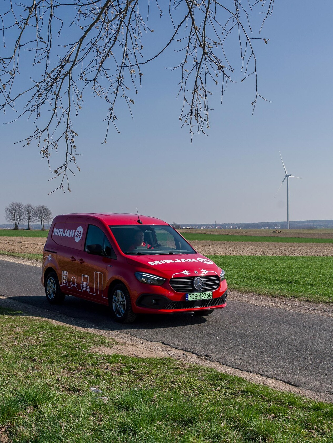 Czerwony eCitany Mercedes-Benz z logiem firmy Mirjan24 na wiejskiej drodze. W tle widać elektrownię wiatrową.