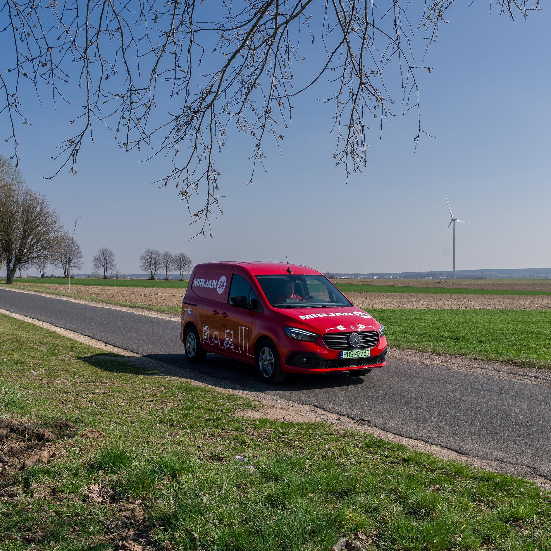 eCitany w firmie Mirjan24: niezawodne wsparcie na każdy dzień | Mercedes-Benz Czerwony eCitany Mercedes-Benz z logiem firmy Mirjan24 na wiejskiej drodze. W tle widać elektrownię wiatrową.