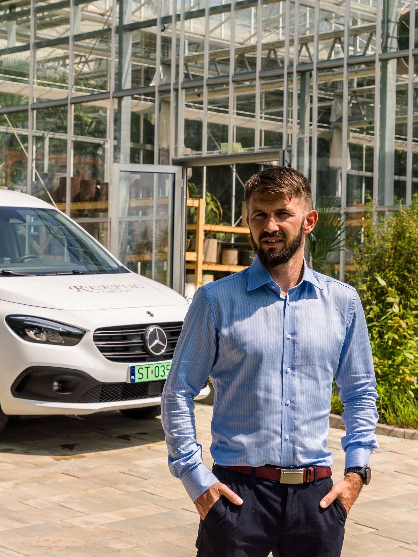 Mężczyzna w niebieskiej koszuli stoi przed białym eCitanem Mercedes-Benz oraz wysoką szklarnią.