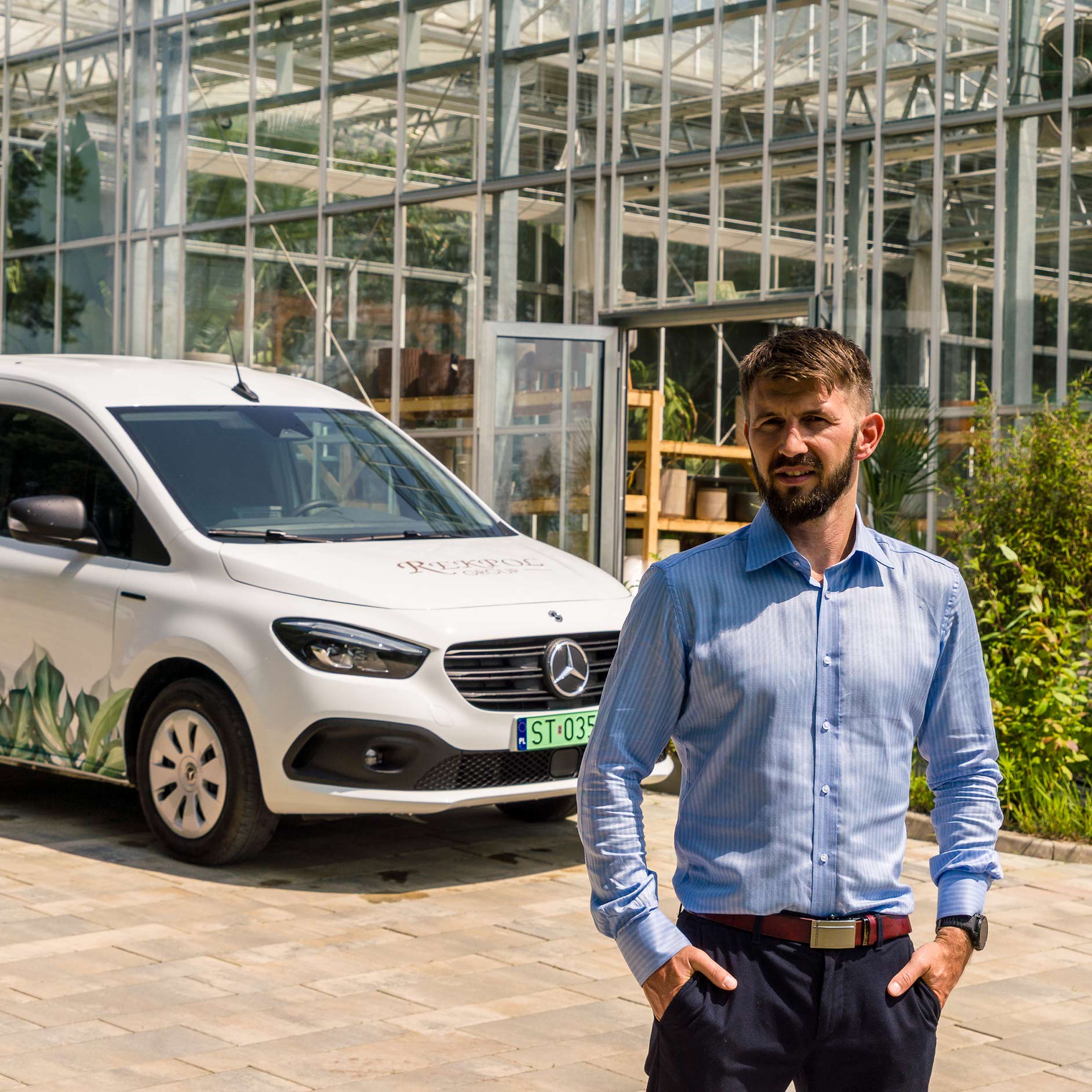 eCitany w Rekpol Group Mężczyzna w niebieskiej koszuli stoi przed białym eCitanem Mercedes-Benz oraz wysoką szklarnią.